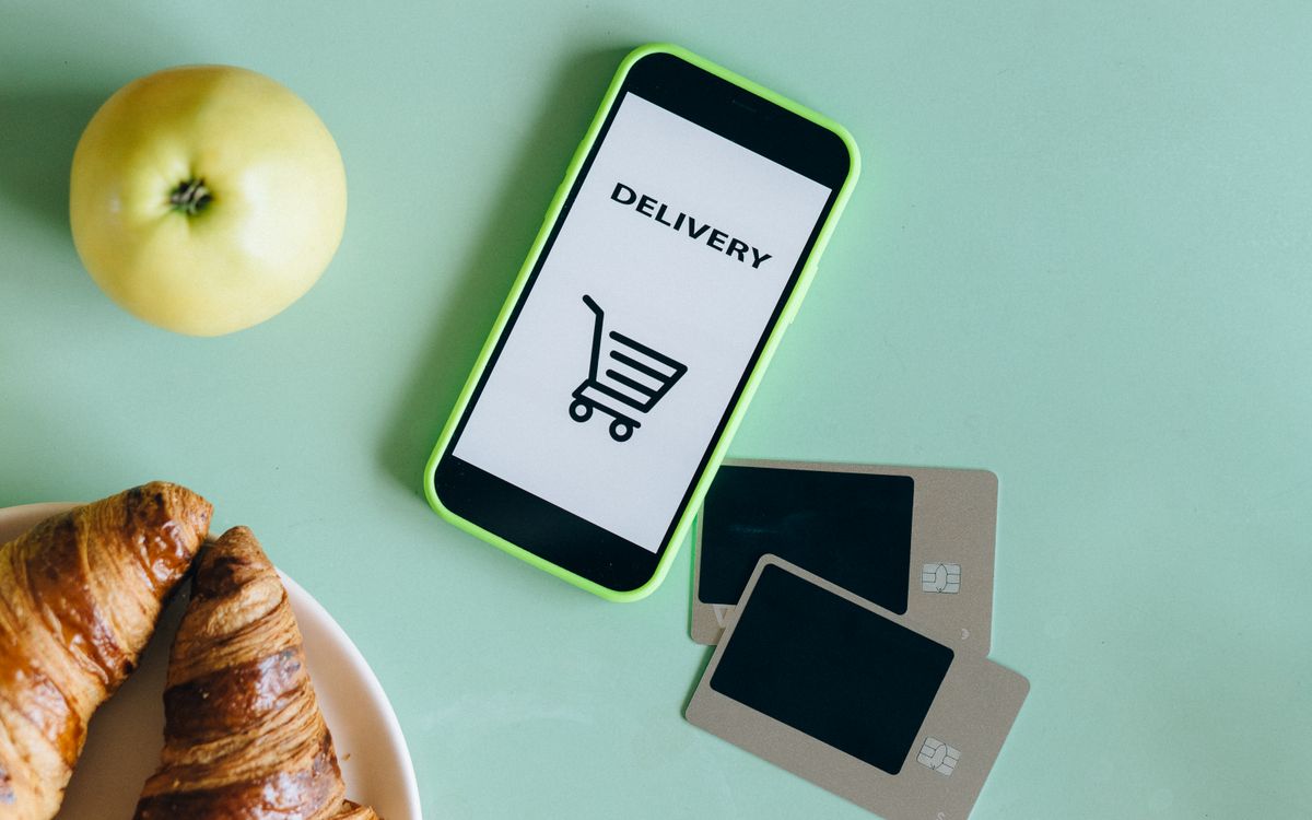 Celular mostrando app de delivery com ícone de carrinho, ao lado de comida e cartões de pagamento