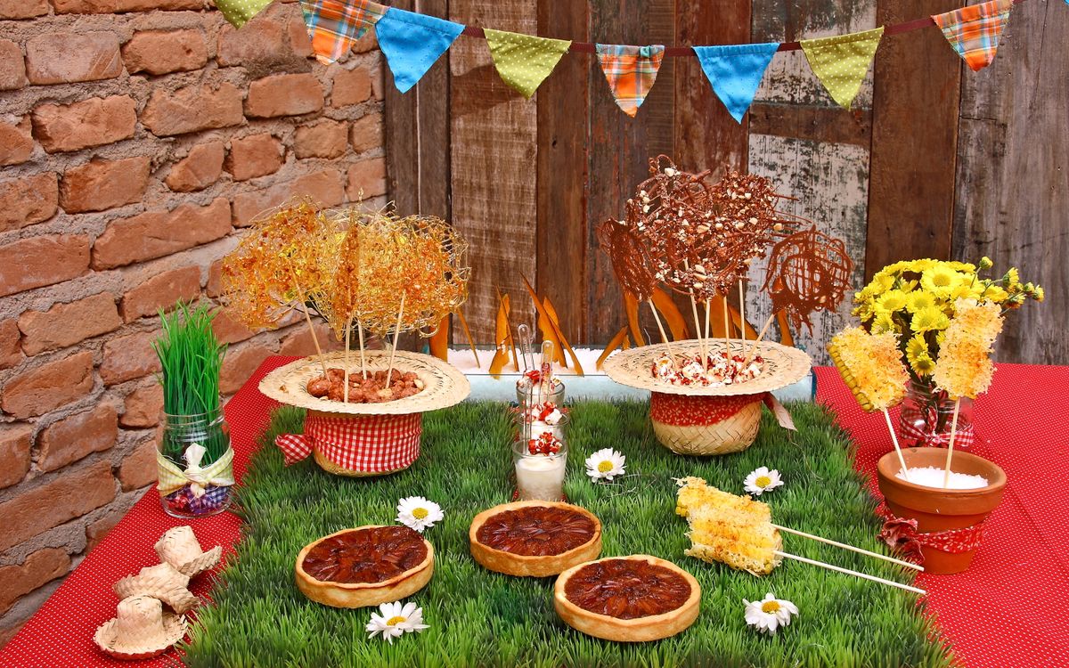 Mesa de festa junina decorada com bandeirinhas coloridas, comidas típicas, flores e palha sobre toalha xadrez