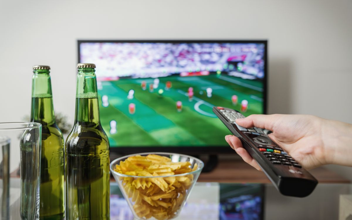 Televisão exibindo uma partida de futebol ao fundo, com cervejas geladas, uma tigela de batatas fritas e uma mão segurando o controle remoto em primeiro plano