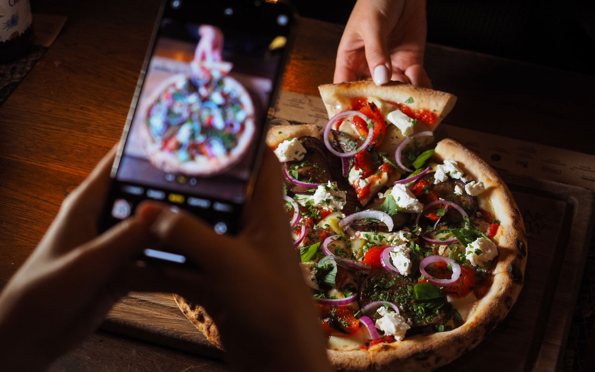 Mãos segurando um celular que fotografa uma pizza com cebola roxa, queijo branco e manjericão sobre tábua de madeira, enquanto outra mão tira uma fatia