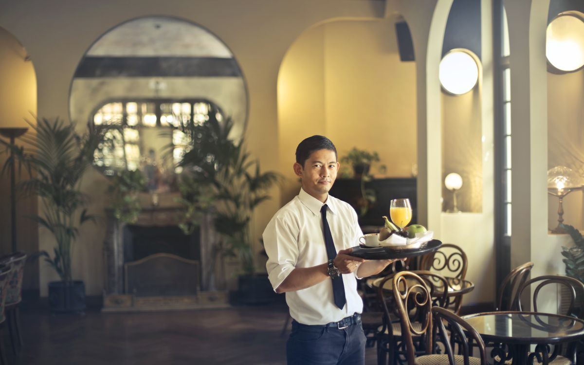 Garçom de camisa branca e gravata segurando uma bandeja com suco de laranja, café e frutas no salão de um restaurante